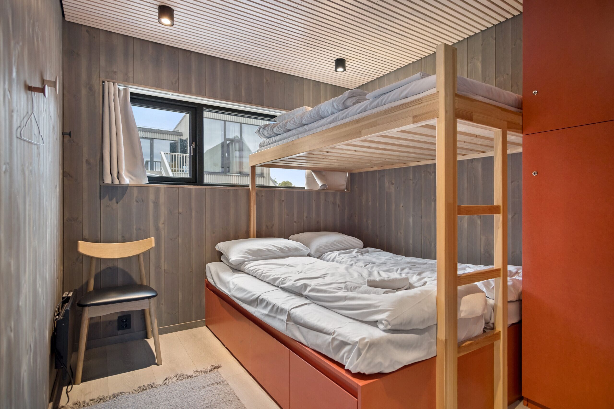 A bunk bed with a wooden frame, a window with curtains, a chair, and a small desk.