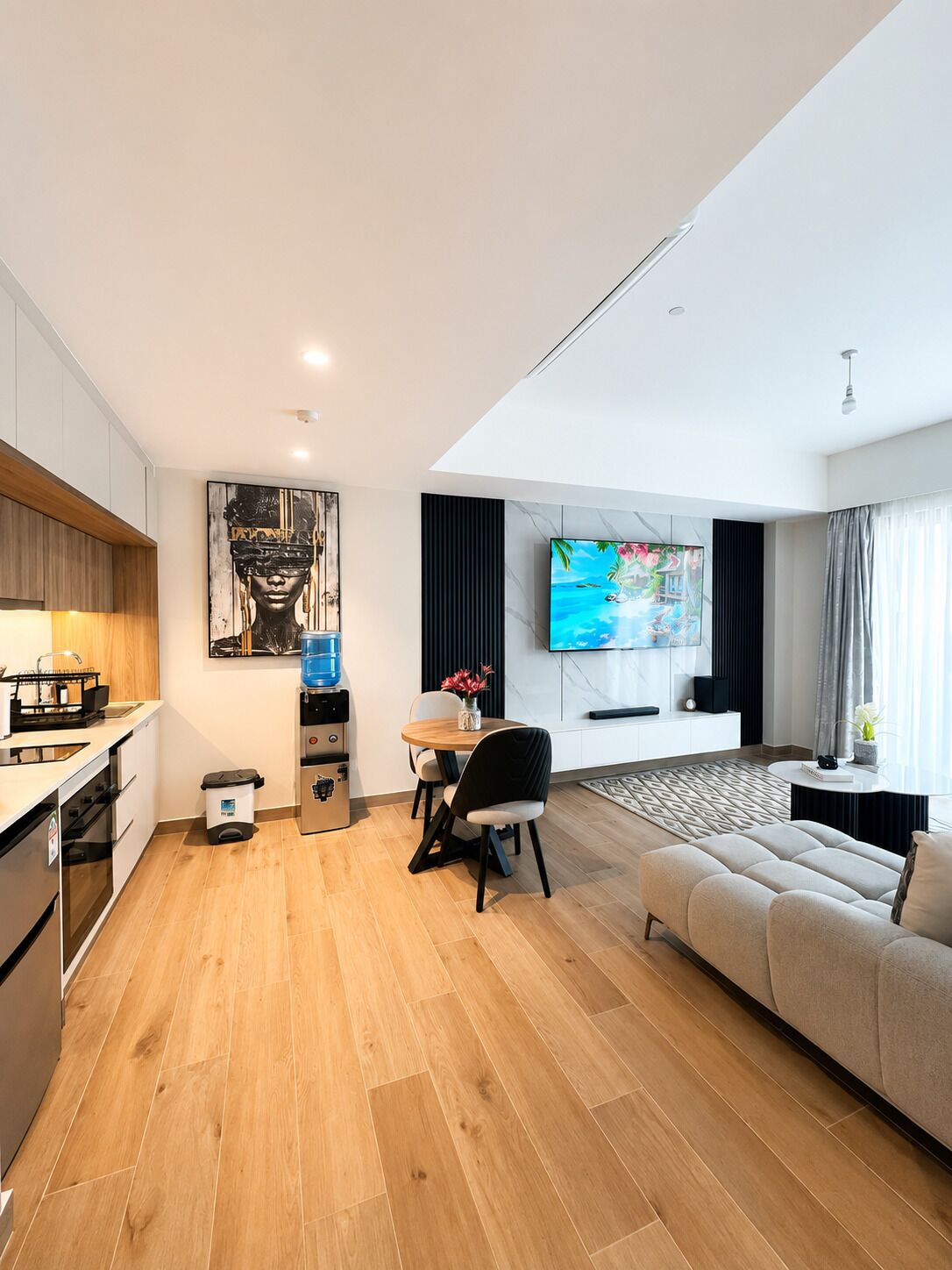 A modern living room with a sofa, a dining table with chairs, a kitchen area with a sink and cabinets, and a flat-screen TV mounted on the wall.