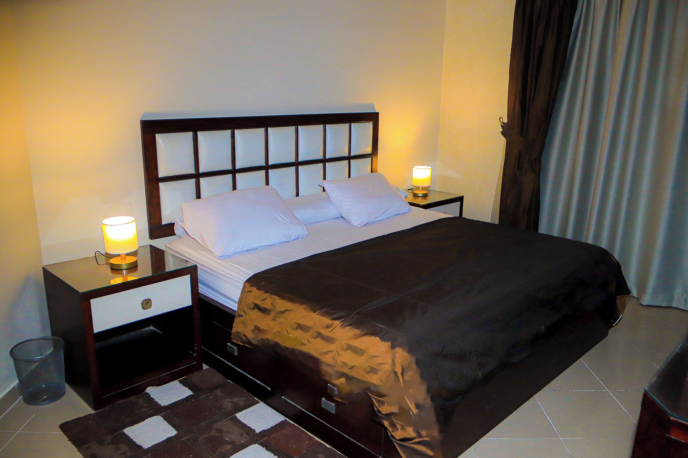 A neatly made bed with a headboard, two bedside tables with lamps, and a window with curtains.