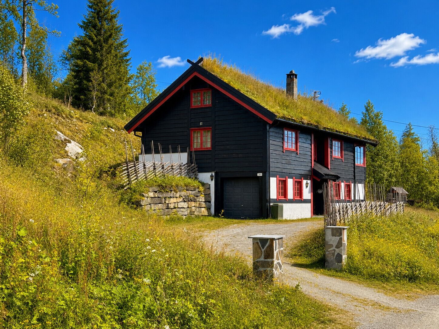 Une maison noire aux ornements rouges et au toit de chaume, entourée de verdure et sous un ciel dégagé.