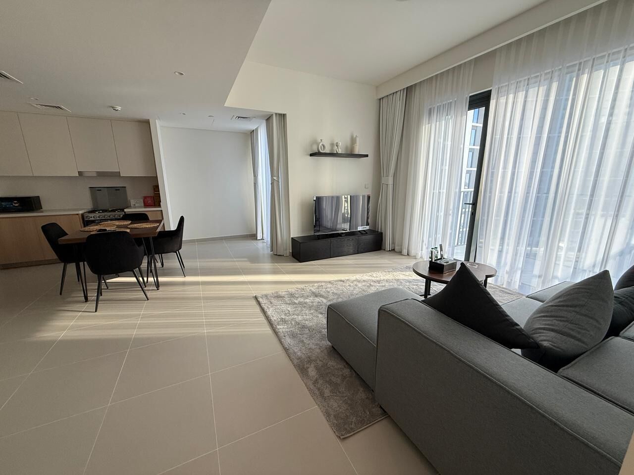 Una sala de estar moderna con un sofá gris, un comedor con mesa y sillas, un televisor de pantalla plana y ventanas amplias con cortinas translúcidas.