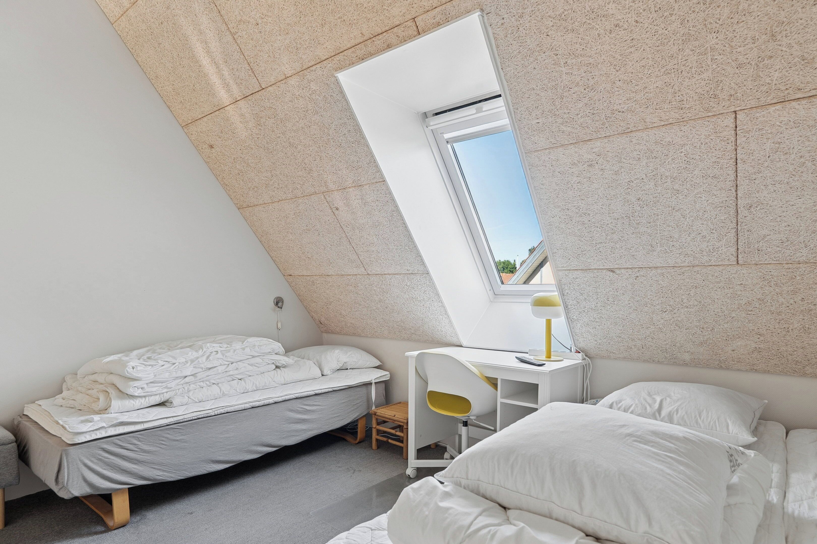 A small, modern bedroom with a slanted ceiling, a single bed, a desk with a chair, and a skylight.