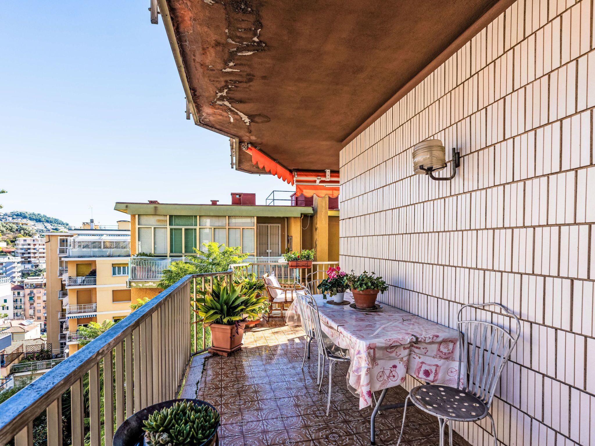 A balcony with a tiled floor, potted plants, a table with chairs, and a view of buildings.