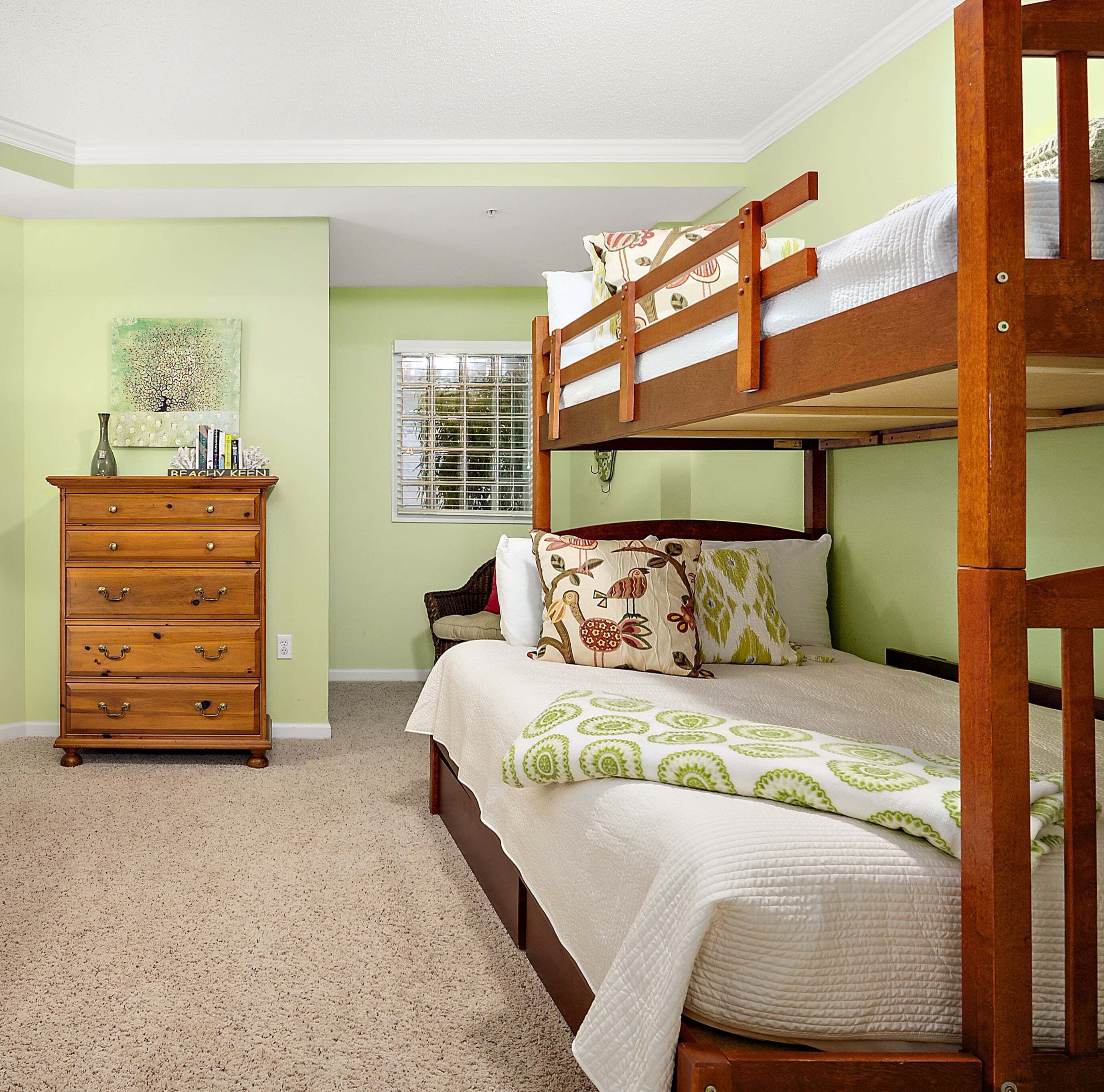 A bunk bed with a wooden frame, a dresser with drawers, a window with blinds, and a wall with a framed picture.