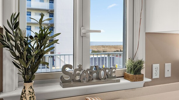 Et vindue med udsigt til en bygning, et dekorativt skilt med teksten 'SEASHORE', en potteplante og et ternet stof på et vindueskar.