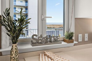 Ein Fenster mit Blick auf ein Gebäude, ein dekoratives Schild mit der Aufschrift 'SEASHORE', eine Topfpflanze und ein kariertes Tuch auf einem Fensterbrett.