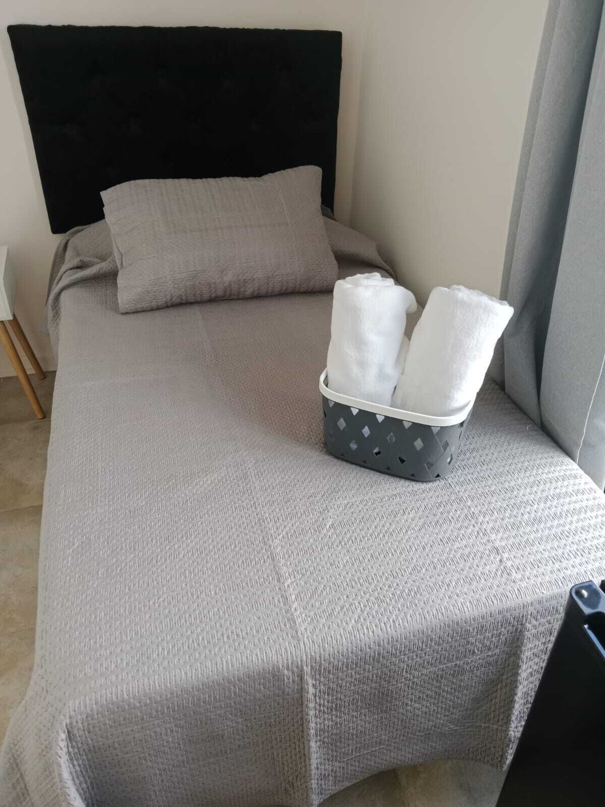 Un lit avec une couvre-lit grise, une tête de lit noire et un oreiller gris. Sur le lit, on trouve un panier gris contenant deux serviettes blanches.