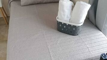 Una cama con una colcha gris, un cabecero negro y una almohada gris. Hay una cesta gris con dos toallas blancas sobre la cama.