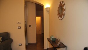 Un pasillo con un sofá, una mesa con adornos, una lámpara en la pared y una puerta que da a otra habitación.