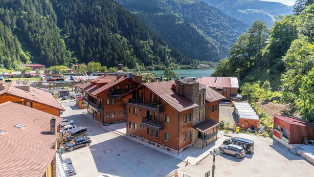 A multi-story building with balconies, a parking area, and a view of a lake surrounded by forested hills. - TURKUAZ APART HOTEL (ÇAYKARA)