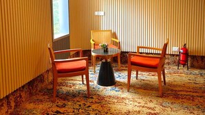 Um quarto com carpete estampado, duas cadeiras de madeira com almofadas, uma mesinha, uma janela e um extintor de incêndio.