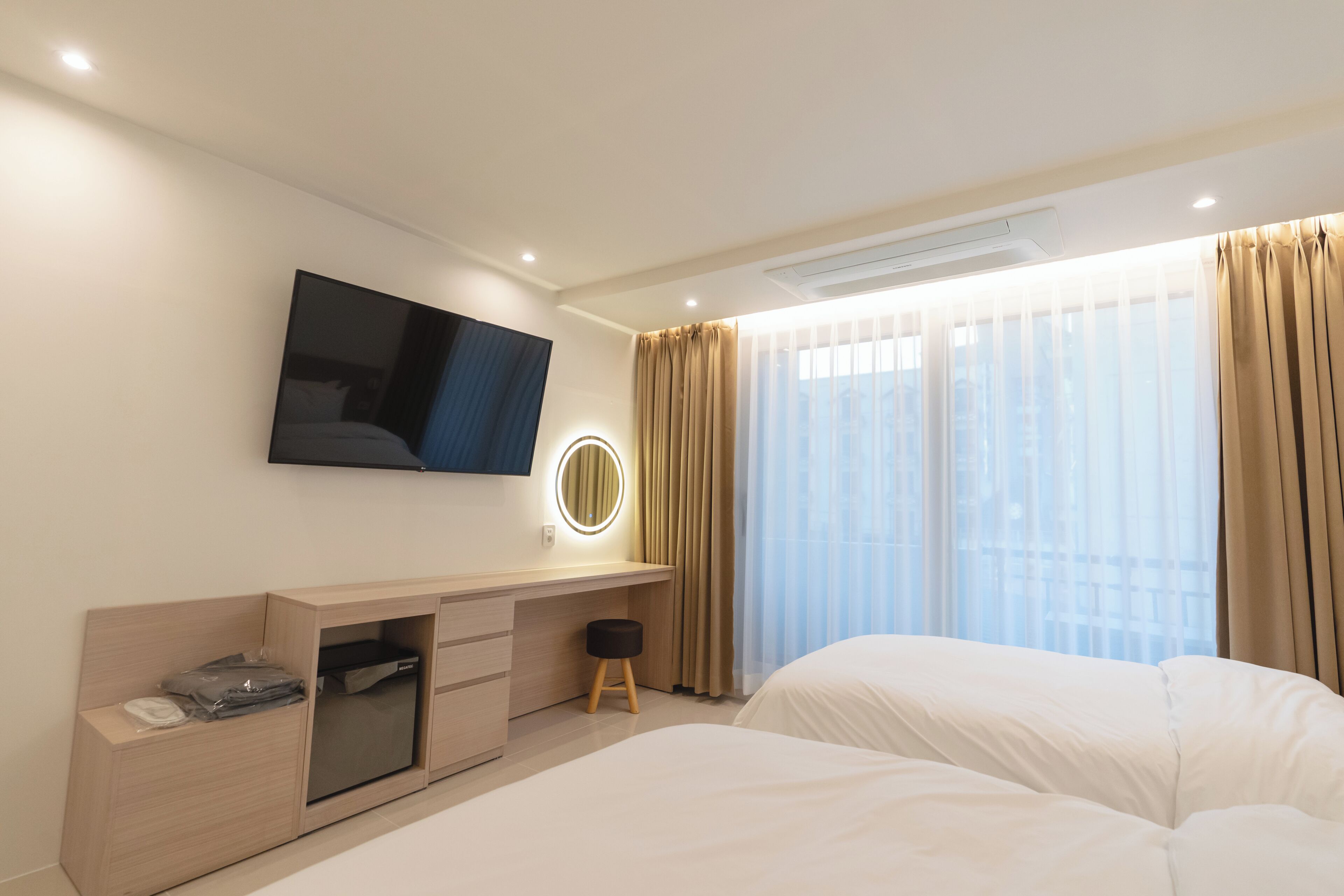 Ein Hotelzimmer mit einem Bett, einem an der Wand befestigten Fernseher, einem Schreibtisch mit Spiegel und einem Fenster mit Vorhängen.
