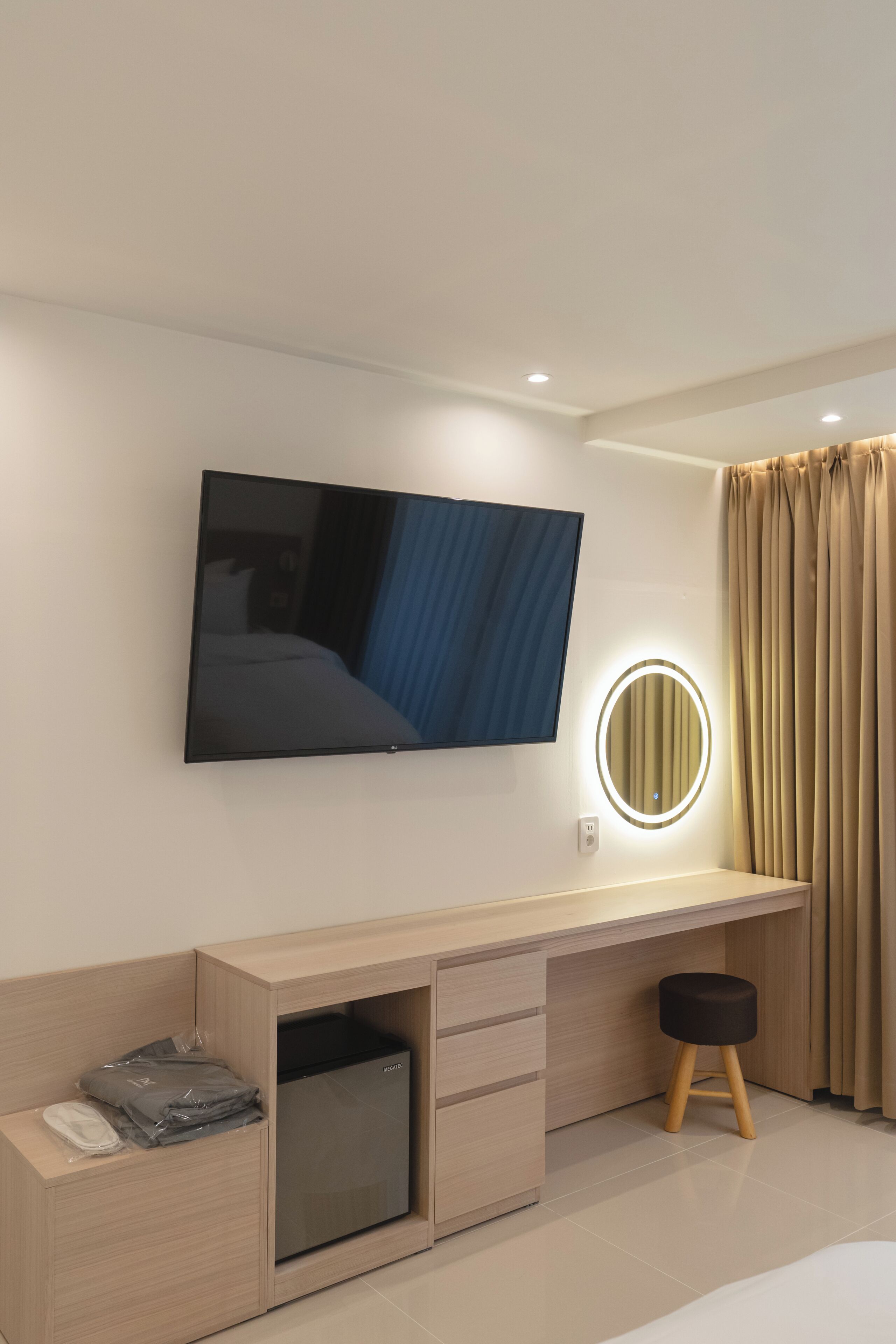 A modern hotel room with a flat-screen TV, a bedside mirror, a desk with drawers, a small stool, and a bed.