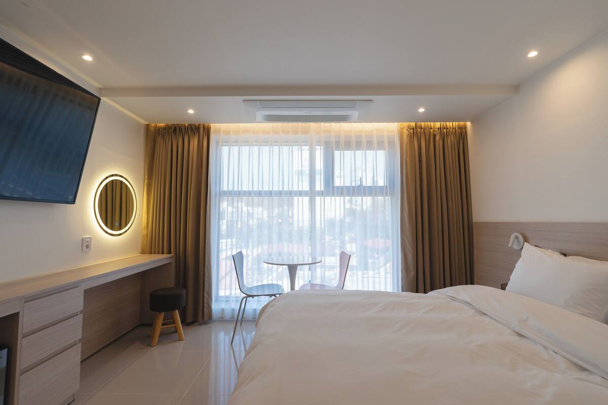 A hotel room with a bed, a desk, a chair, a round mirror, and a window with curtains.