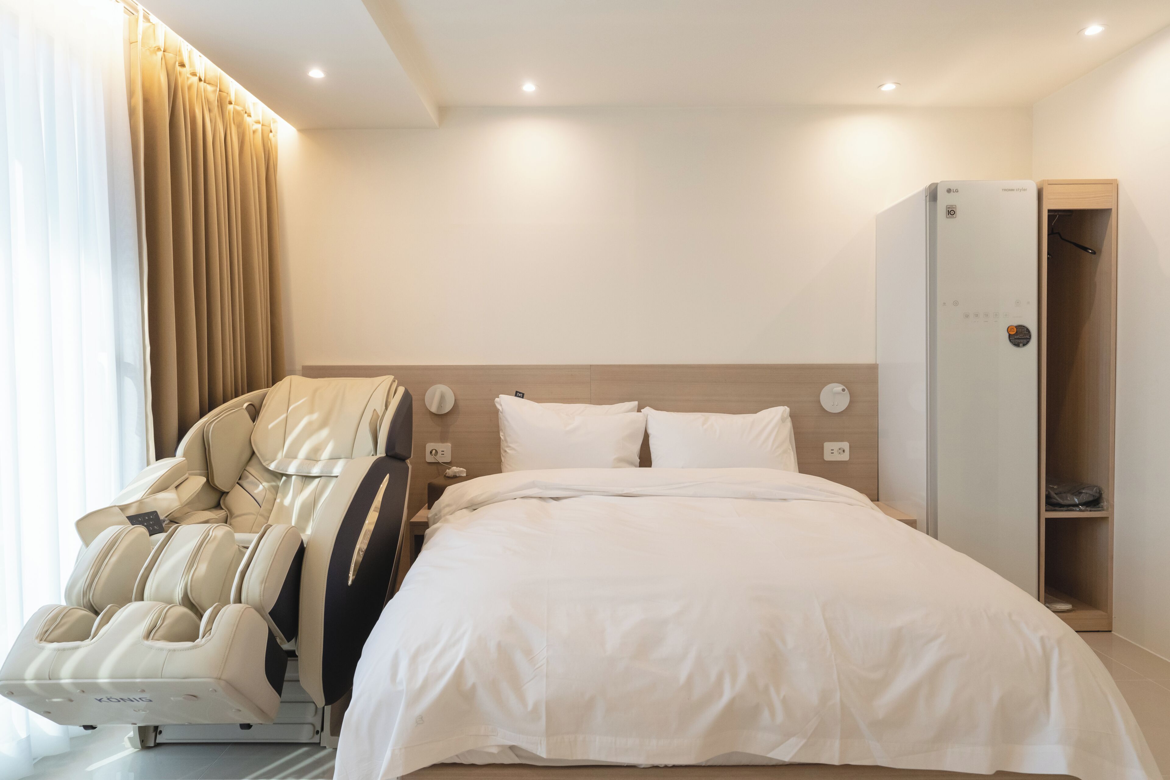 Habitación de hotel con sillón de masajes, cama con sábanas blancas, cabecero de madera y armario empotrado.