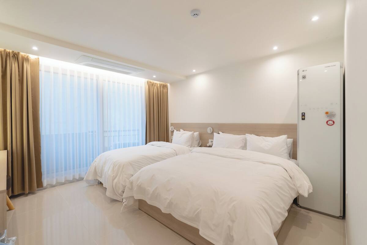 A hotel room with two beds, a large window with curtains, a wall-mounted air conditioner, and a door with a lock.