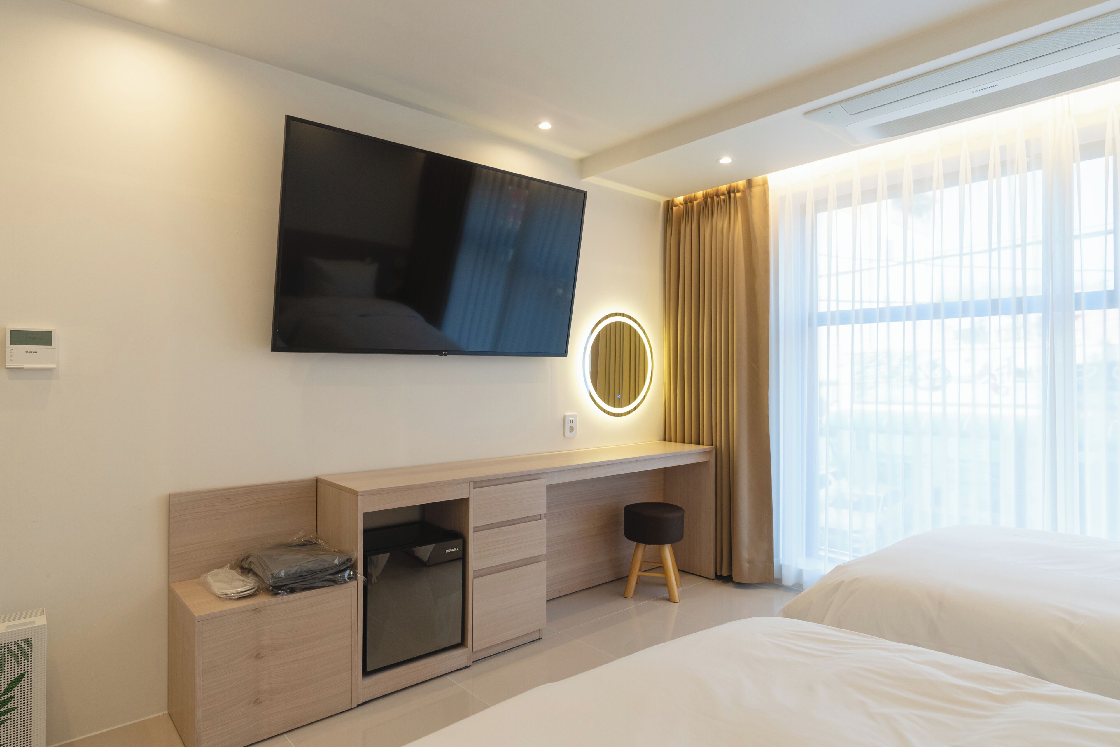 Una habitación de hotel moderna con televisor de pantalla plana, una mesita de noche con lámpara, un ventanal con cortinas y una cama.