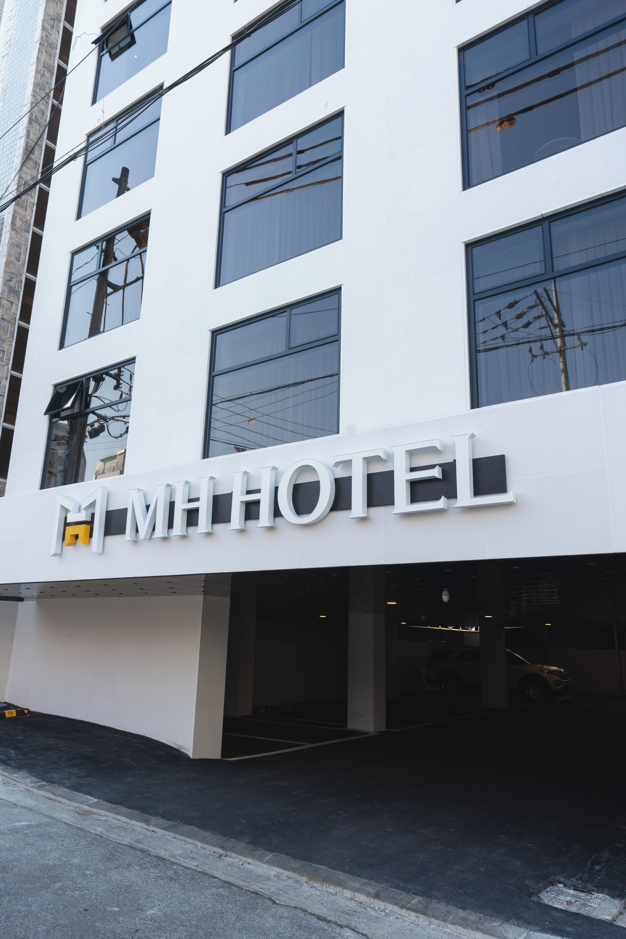 Ein modernes Hotelgebäude mit einem auffälligen 'MH HOTEL'-Schild, zahlreichen Fenstern und einem Parkplatz.