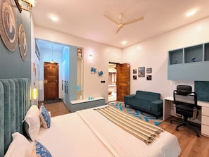 Ein modernes Hotelzimmer mit Bett, Schreibtisch, Sessel, Sofa, Deckenventilator und einem durchgehenden Türspalt sichtbaren Badezimmer.