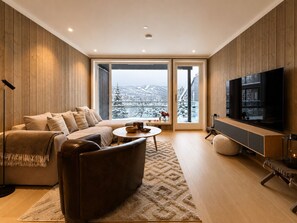 Ein modernes Wohnzimmer mit Sofa, Couchtisch und Fernseher, mit Holzwänden und einem großen Fenster mit Schneeaussicht.