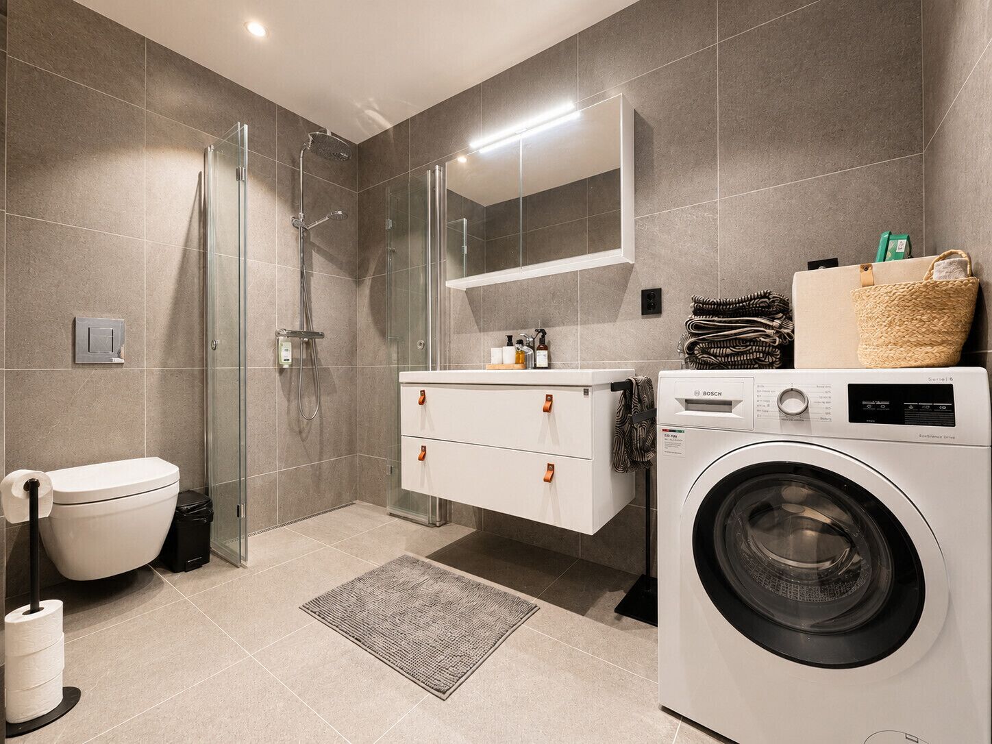 A modern bathroom with a glass shower enclosure, a Bosch washing machine, a toilet, and a mirror above the sink.