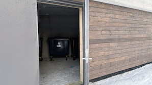 Une porte de garage ouverte, avec une benne à ordures à l’intérieur, un sol en béton et un mur extérieur en bois.
