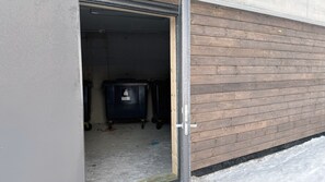 Eine offene Garagentür, ein Müllcontainer im Inneren, ein Betonboden und eine Holzfassade.
