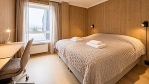 Une chambre à coucher avec un grand lit, un bureau avec une chaise, une fenêtre avec des rideaux et un luminaire fixé au mur.