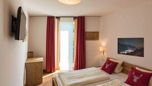 Ein Schlafzimmer mit Bett, an der Wand montiertem Fernseher, Nachttischchen aus Holz, einer Nachttischlampe, einer Deckenleuchte und einem Fenster mit roten Vorhängen.