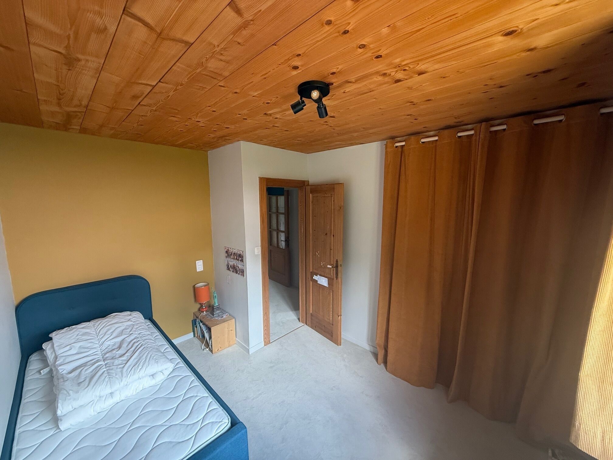 Un dormitori amb sostre de fusta, un llit, una petita taula de nit amb una làmpada i una porta que condueix a una altra habitació.