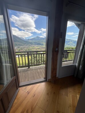 A room with a wooden floor, a sliding glass door leading to a balcony with a view of mountains and a town. - Chalet Mont Joly, mountain views, 20 minutes from Chamonix-Mont-Blanc (Passy)