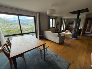 A spacious living area with a large wooden table, a sofa, and a view of a mountainous landscape through large windows. - Chalet Mont Joly, mountain views, 20 minutes from Chamonix-Mont-Blanc (Passy)