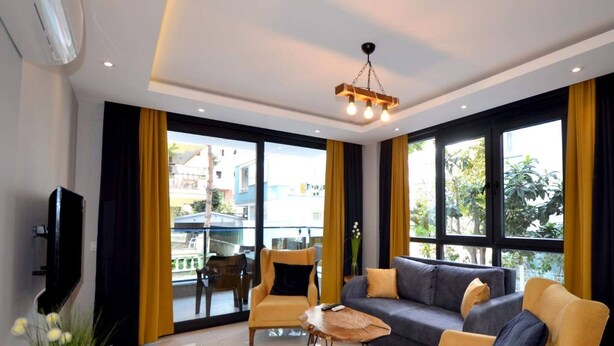 Una sala de estar moderna con un sofá, sillones, una mesa de centro y un televisor. Amplias ventanas con cortinas permiten la entrada de luz natural y ofrecen vistas al exterior.