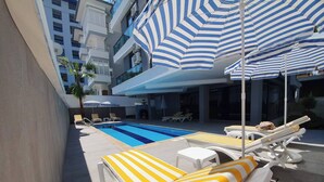 Zona de piscina con sillas reclinables y sombrillas, una piscina e un edificio moderno al fondo.