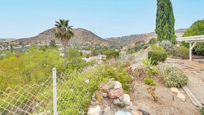 Ein von einer Hecke umgebener Garten mit verschiedenen Pflanzen und einem hohen Baum, mit Blick auf eine hügelige Landschaft mit Häusern.