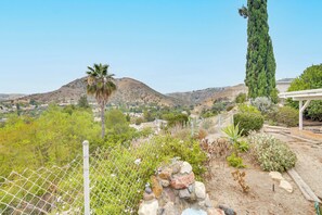 Ein von einer Hecke umgebener Garten mit verschiedenen Pflanzen und einem hohen Baum, mit Blick auf eine hügelige Landschaft mit Häusern.