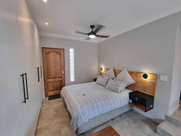 A bedroom with a bed, a ceiling fan, a nightstand, a wooden headboard, and a door. - Saxon Guest Cottage Suite (Cape Town)