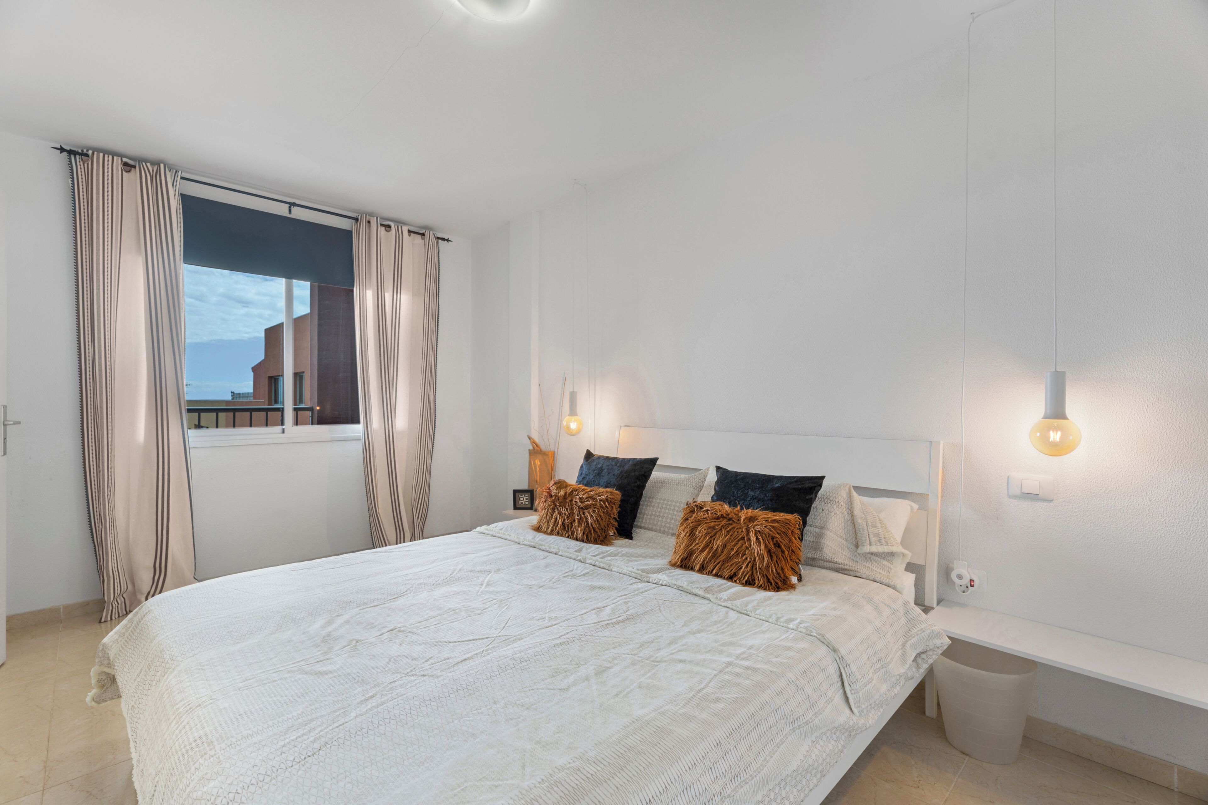 A modern bedroom with a large bed, two bedside tables, a nightstand, and a window with curtains.