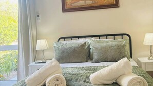 Un dormitorio con cama, almohadas, un cuadro, una ventana con cortinas y una lámpara de noche.