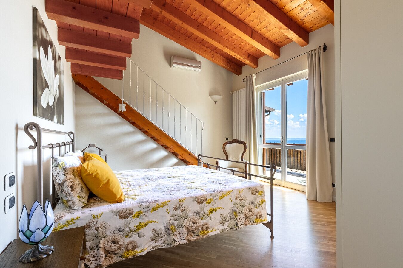A bedroom with a bed, a staircase, a window with curtains, and a view of the sea.