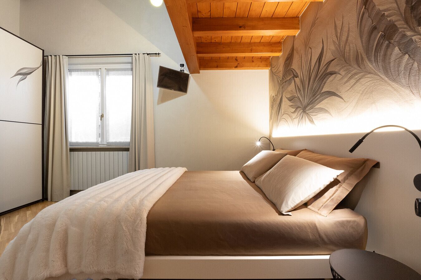 A bedroom with a bed, a TV mounted on the wall, a window with curtains, and a wooden ceiling.
