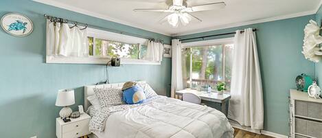 A bedroom with a bed, bedside table, nightstand, ceiling fan, window with curtains, and a desk with a chair.