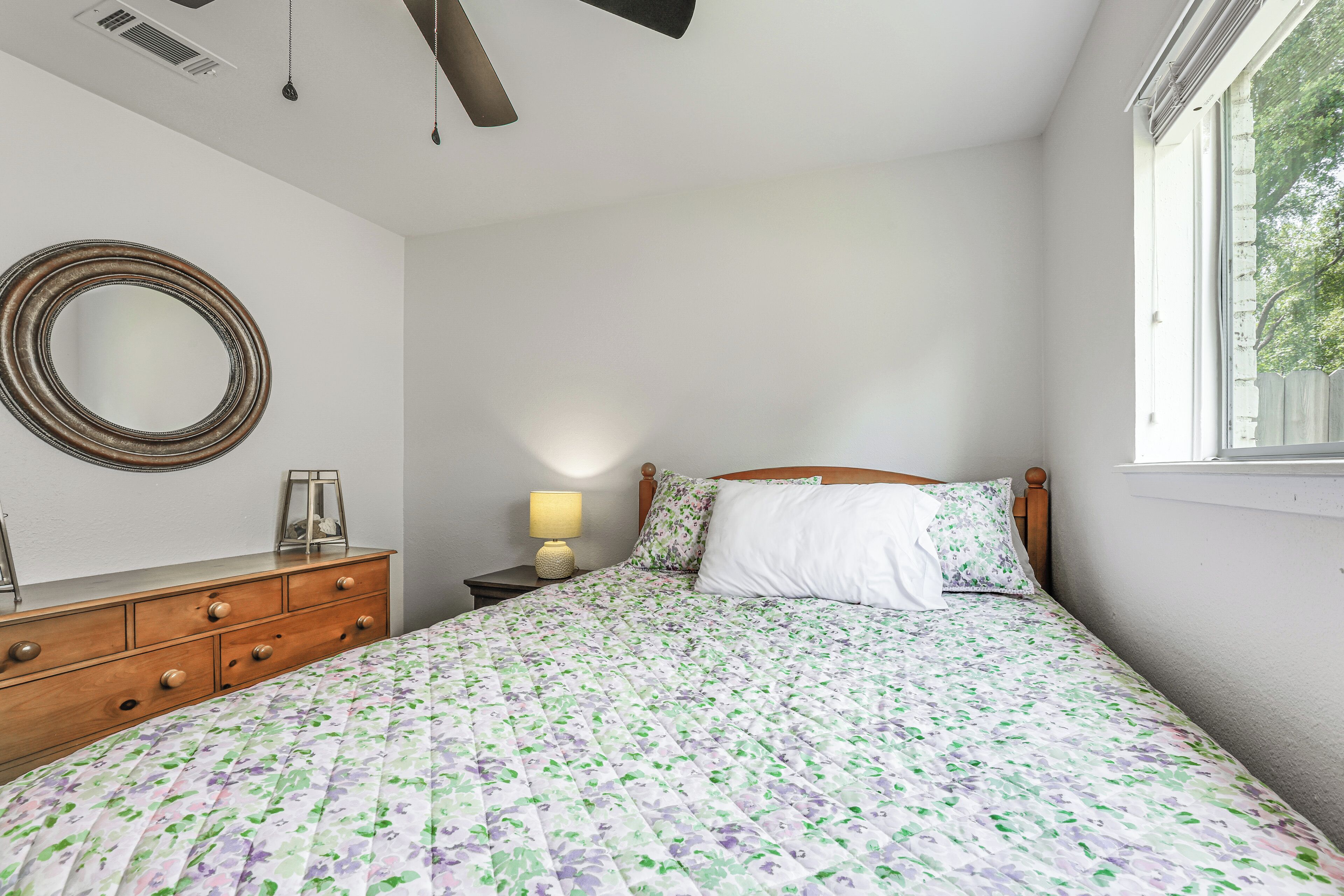 Une chambre à coucher avec un lit, une commode, un miroir, une lampe et une fenêtre.