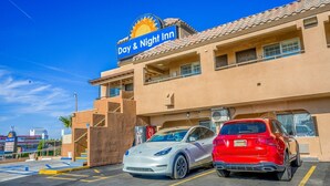 Un hôtel ouvert jour et nuit, avec un parking, deux voitures garées et un ciel bleu dégagé.