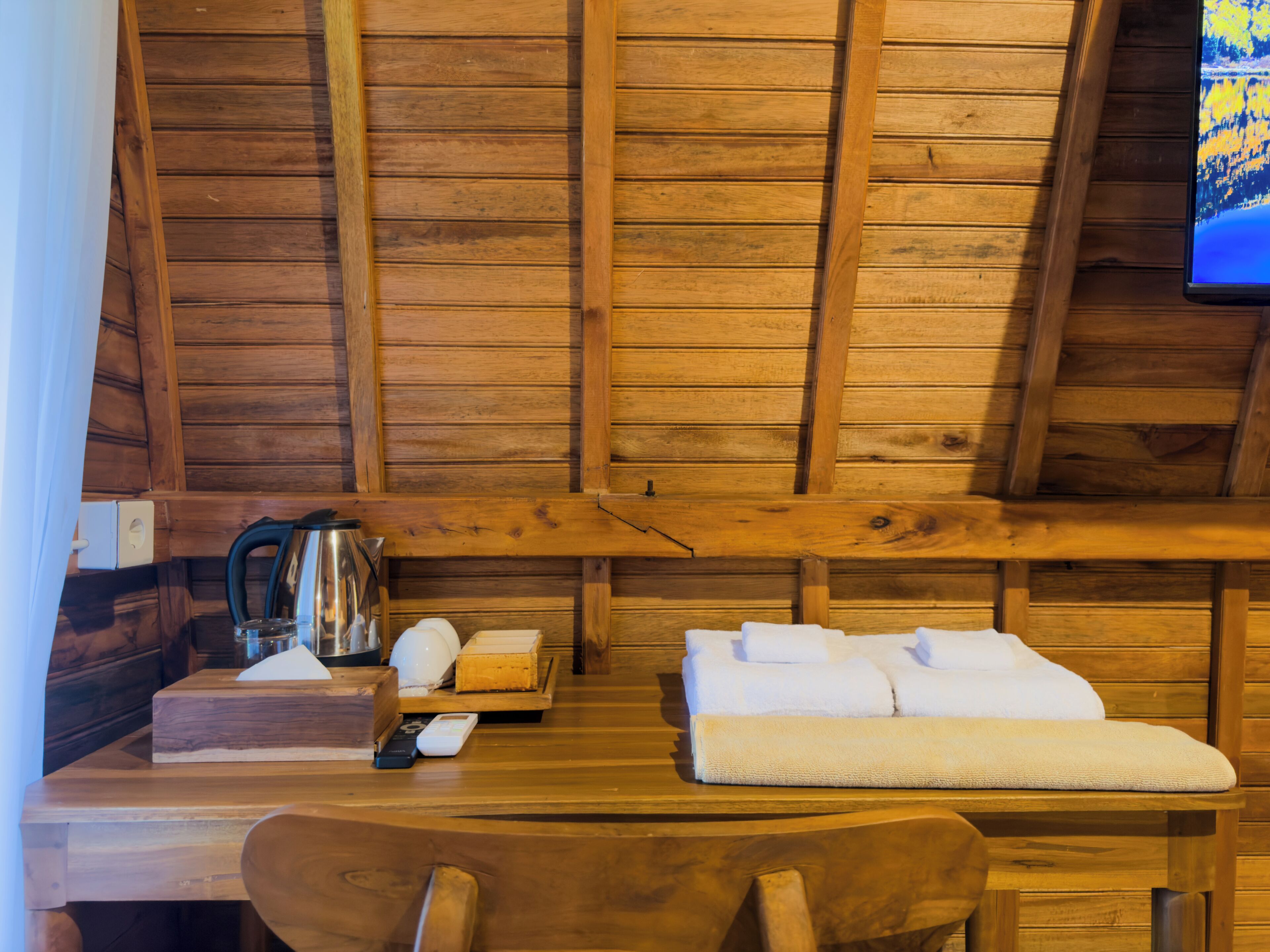 A wooden cabin interior with a wooden table, a sofa, a chair, a kettle, and a television mounted on the wall.