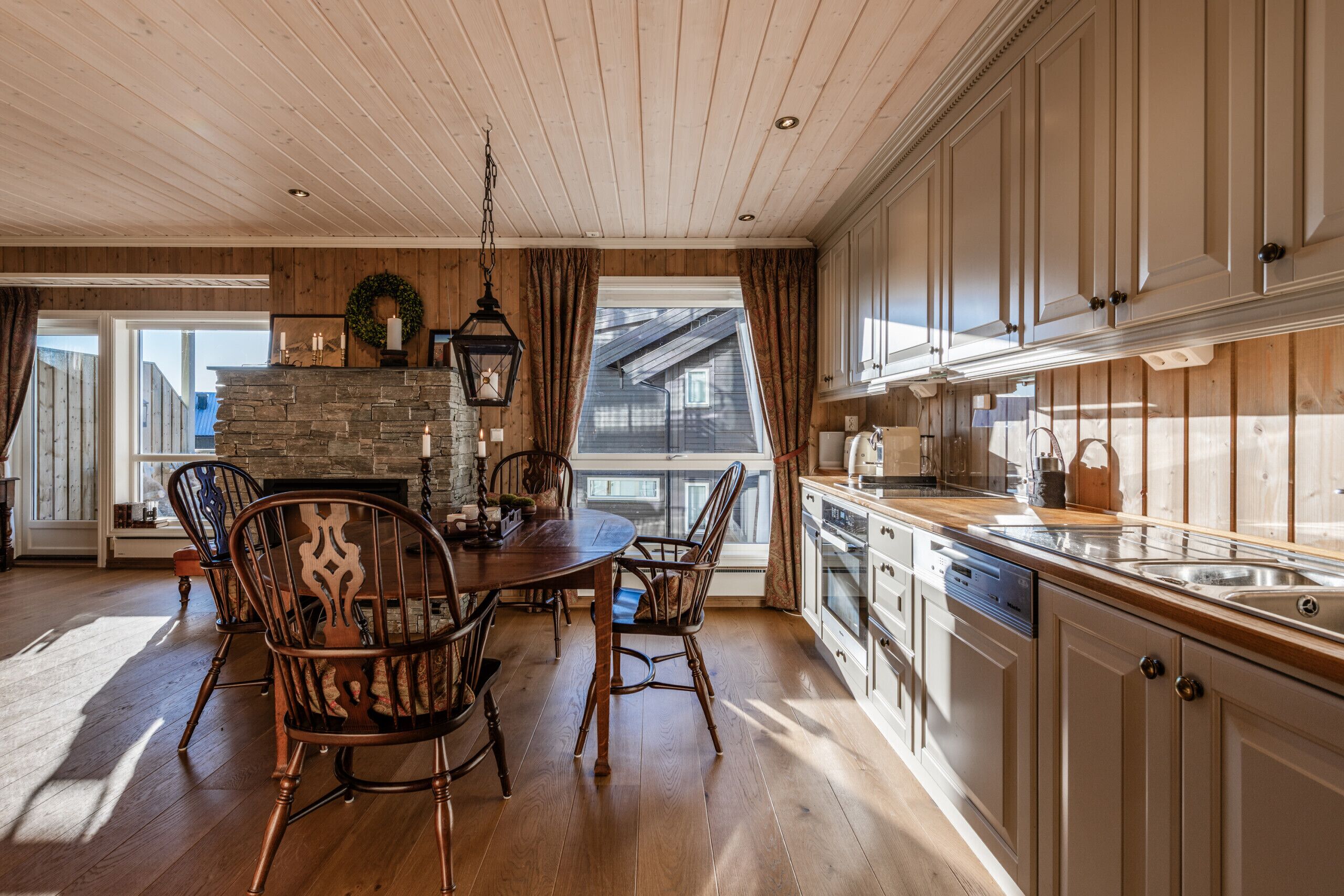 A dining area with a stone fireplace, wooden ceiling, and a view of a house through a window.