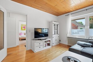 Ein modernes Wohnzimmer mit einem Flachbildfernseher, einem weißen Schrank, einer Couch und einem Fenster mit Blick auf ein Haus.