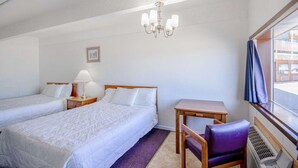 A hotel room with two beds, a desk, a chair, a table, a chandelier, a picture on the wall, and a window with curtains.