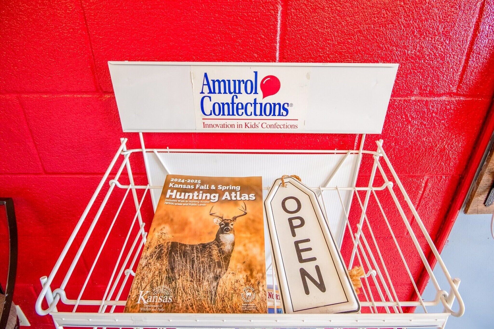 Un présentoir en métal blanc sur lequel est apposée une enseigne indiquant « Amurol Confections », ainsi qu’un livre intitulé « Kansas Fall & Spring Hunting Atlas », accompagné d’une enseigne indiquant « OUVERT ».