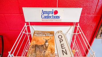 Un présentoir en métal blanc sur lequel est apposée une enseigne indiquant « Amurol Confections », ainsi qu’un livre intitulé « Kansas Fall & Spring Hunting Atlas », accompagné d’une enseigne indiquant « OUVERT ».